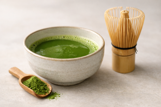 Bowl of prepared matcha with bamboo whisk and matcha powder on a scoop.